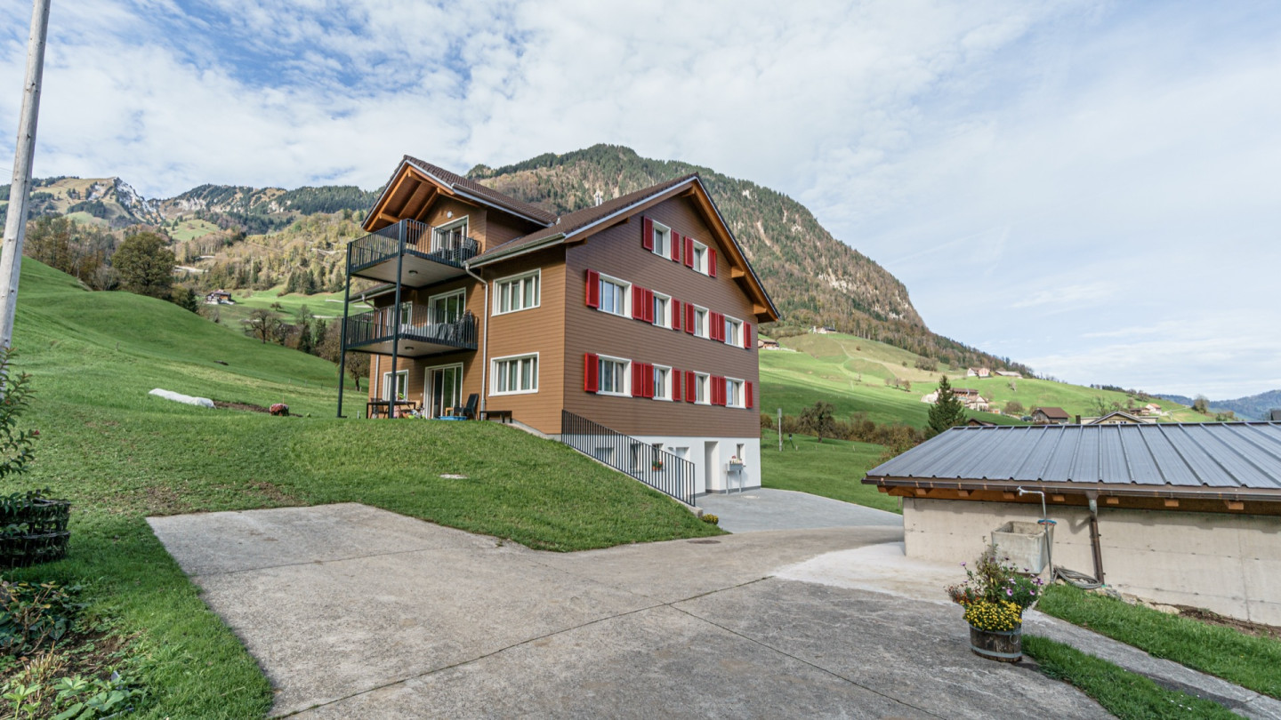 Giebelfassade mit rot gerahmten Fensterläden vor Bergkulisse